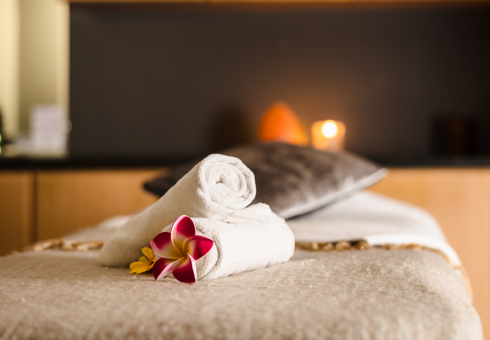 Rolled white towels and a pink flower on a massage table with a pillow. A lit candle is visible in the blurred background.