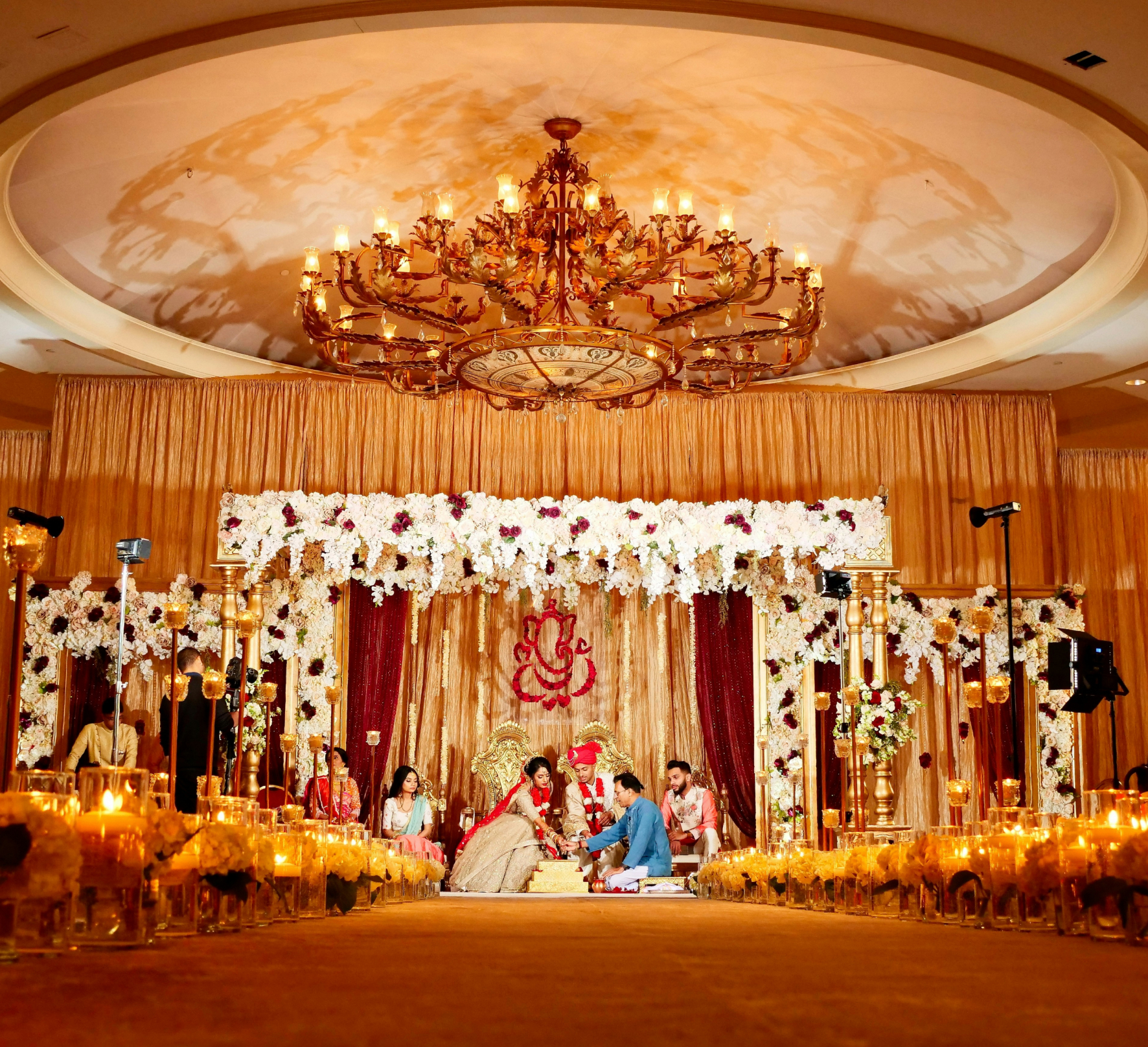 A couple participates in a traditional wedding ceremony under a large chandelier, surrounded by floral decorations and guests on an elaborately decorated stage.