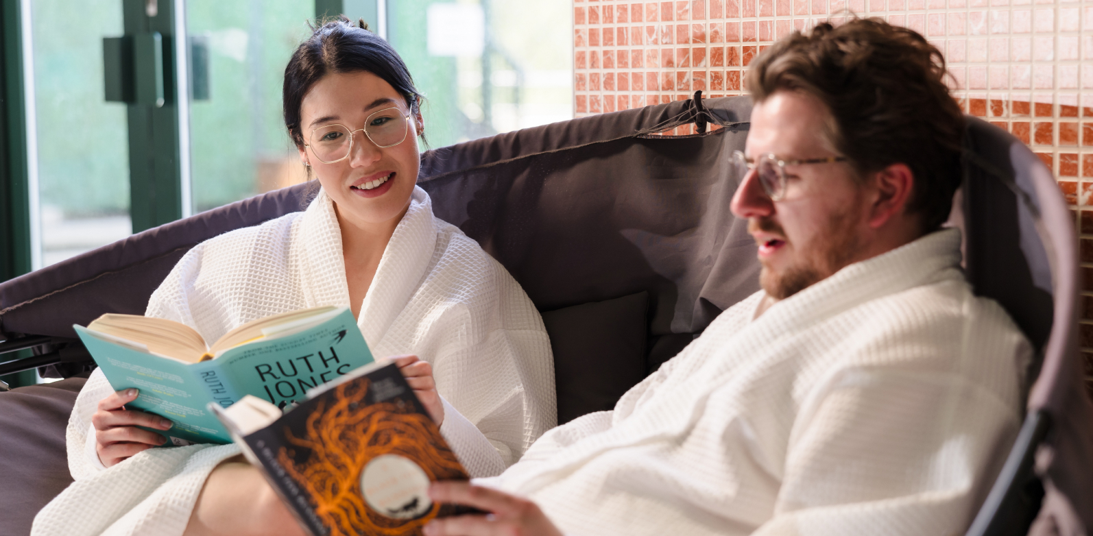 Two people in white robes sit on a sofa, each reading a different book. The woman is smiling while reading.