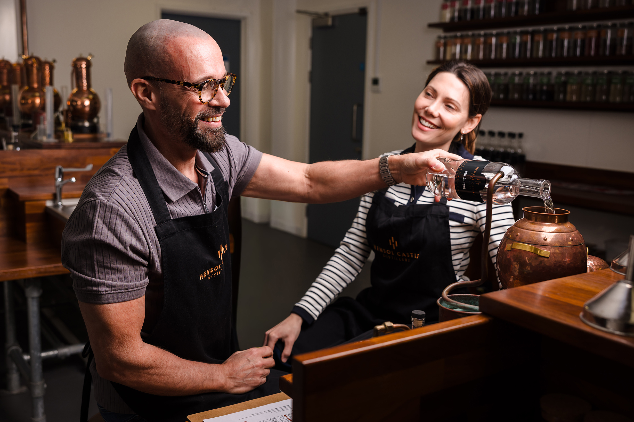 Two people in aprons smile as one pours liquid into a copper still during a gin tour breaks experience.