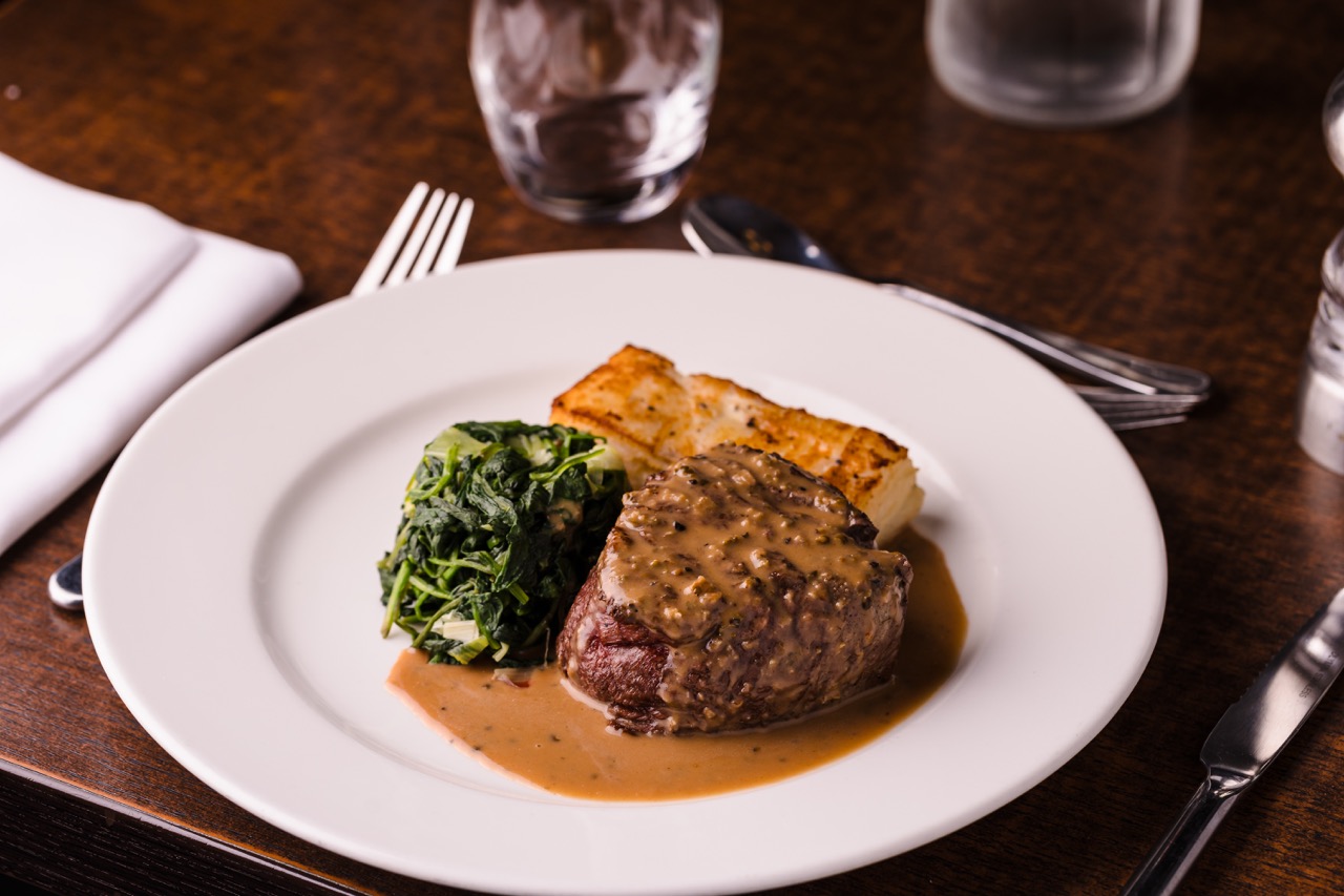 A plate with steak topped with sauce, spinach, and a potato gratin, placed on a wooden table with a fork, knife, glass, and napkin.