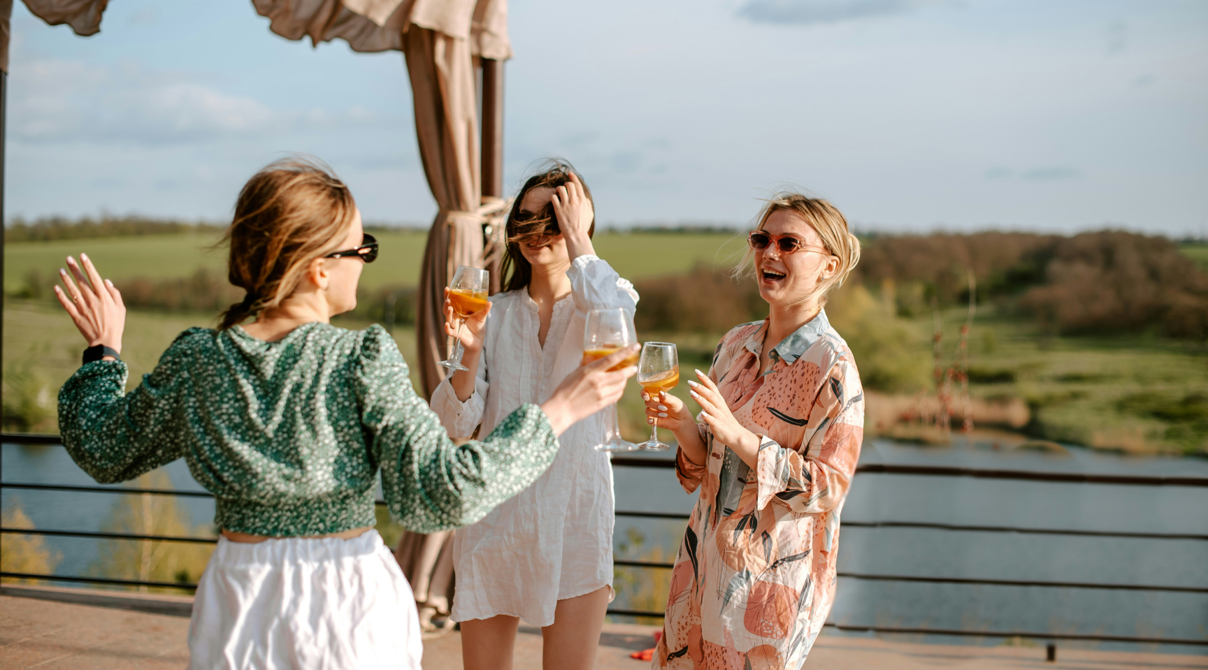 Three people stand on a balcony with drinks in hand, laughing and wearing casual summer clothing with a scenic landscape in the background.