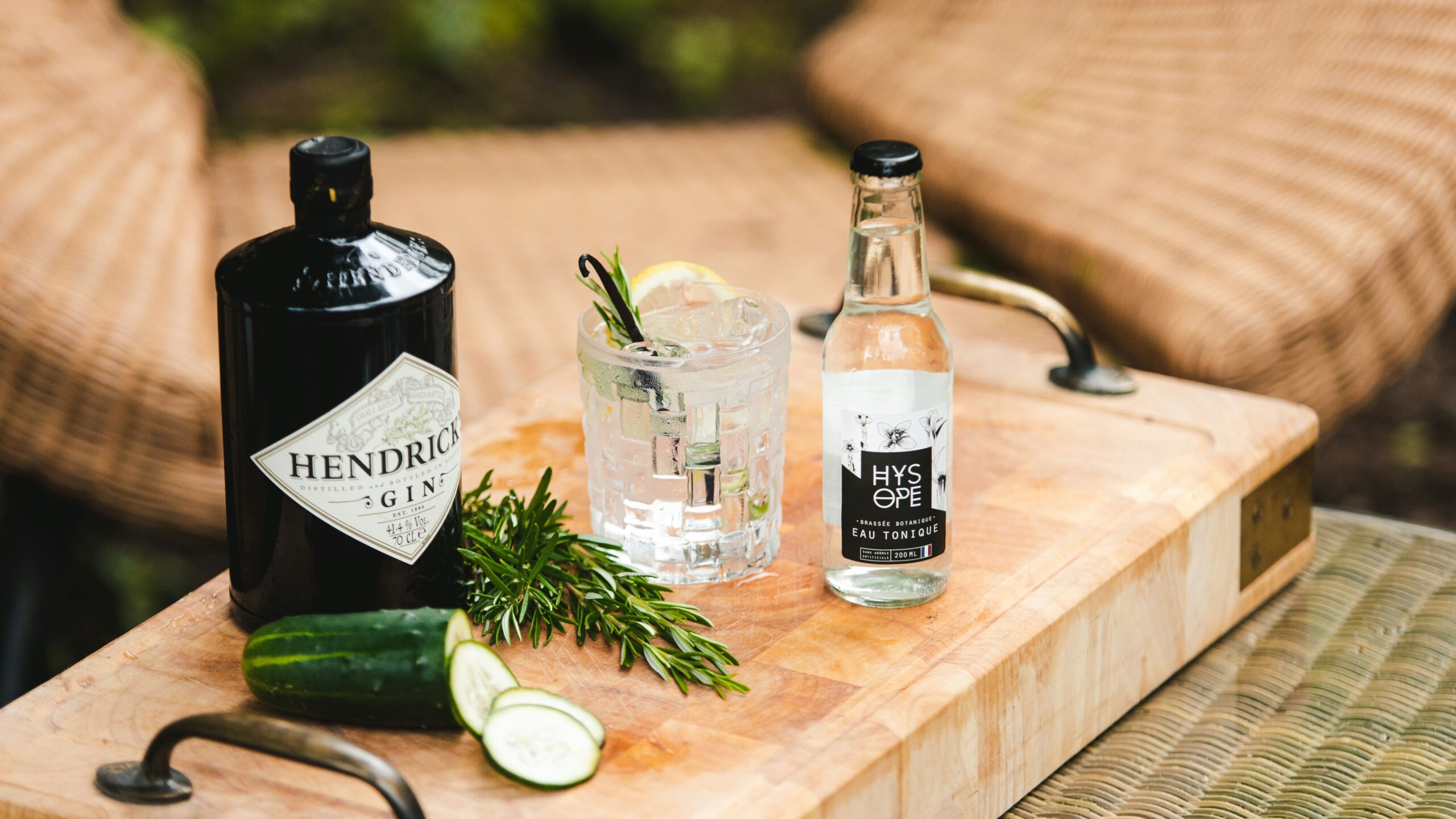 A bottle of Hendrick's Gin, a bottle of tonic water, a glass with a lime garnish, rosemary, and cucumber slices on a wooden tray, with wicker chairs in the background.