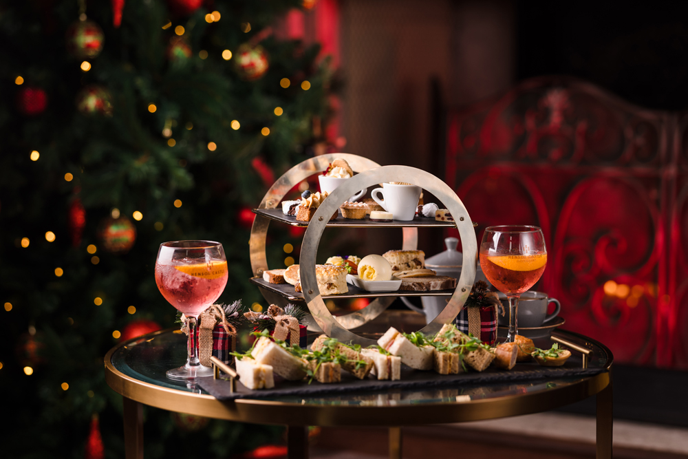 A festive afternoon tea set with sandwiches, pastries, and drinks on a round table in front of a decorated Christmas tree.