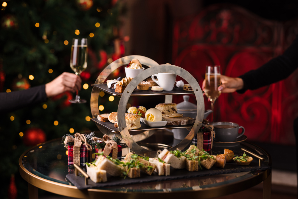 Two people toast with champagne next to a tiered tray of assorted pastries, sandwiches, and desserts on a table.