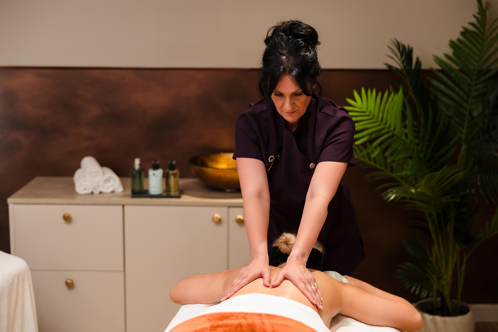 A massage therapist performs a back massage on a client in a spa room with towels, plants, and massage oils in the background.