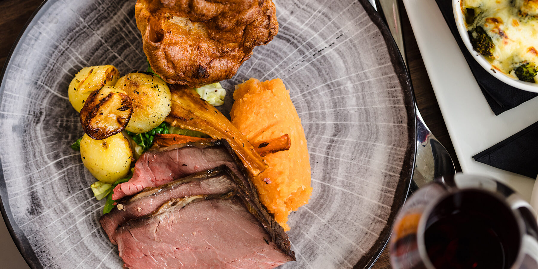 A plate with roast beef slices, Yorkshire pudding, roasted potatoes, carrot mash, and vegetables. A dish of broccoli gratin is partially visible.