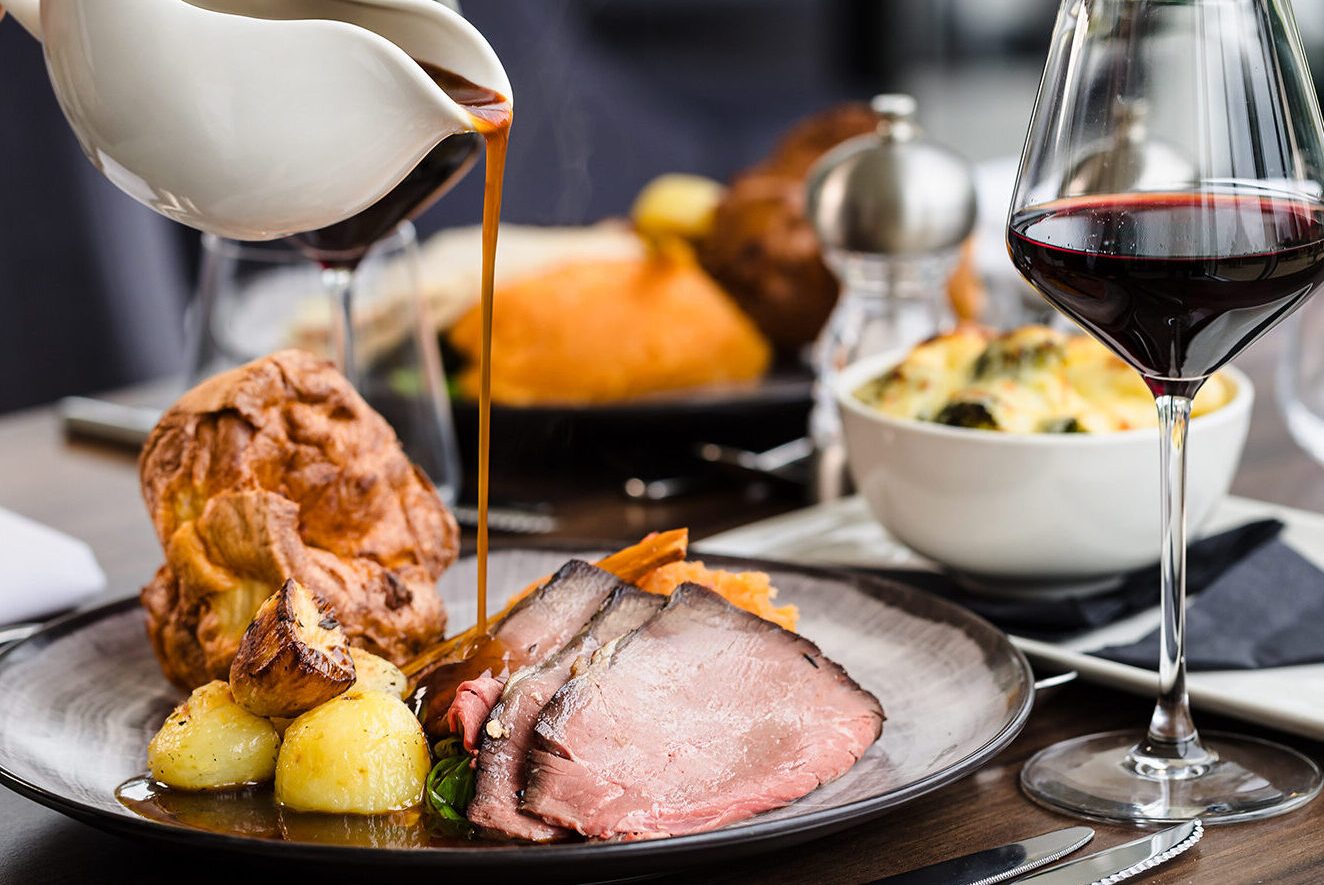 A plate of roast beef, potatoes, and Yorkshire pudding is being drizzled with gravy. A glass of red wine and a bowl of side dish are on the table.