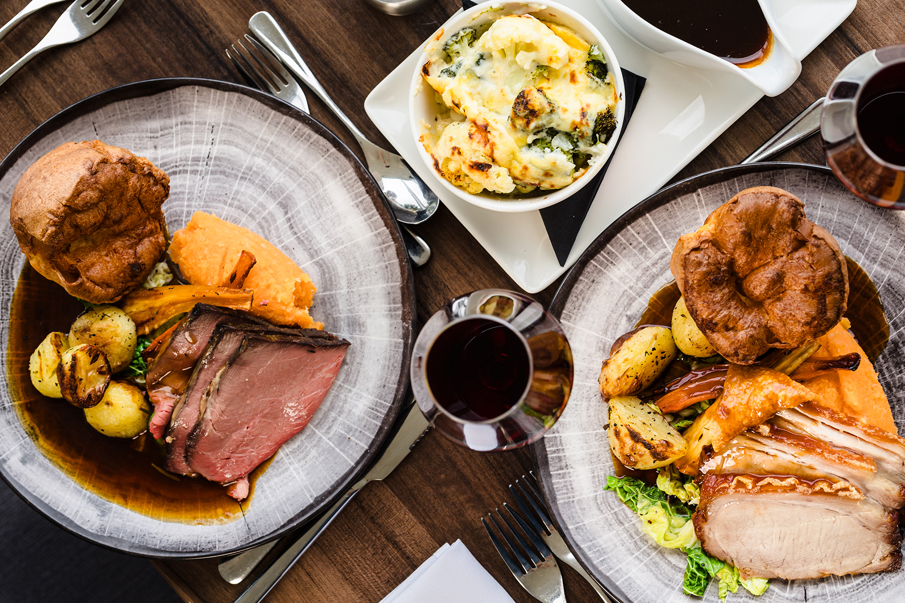Two plates of roast dinner with vegetables, Yorkshire pudding, and gravy. A side dish of creamy spinach and glasses of red wine are on a wooden table.