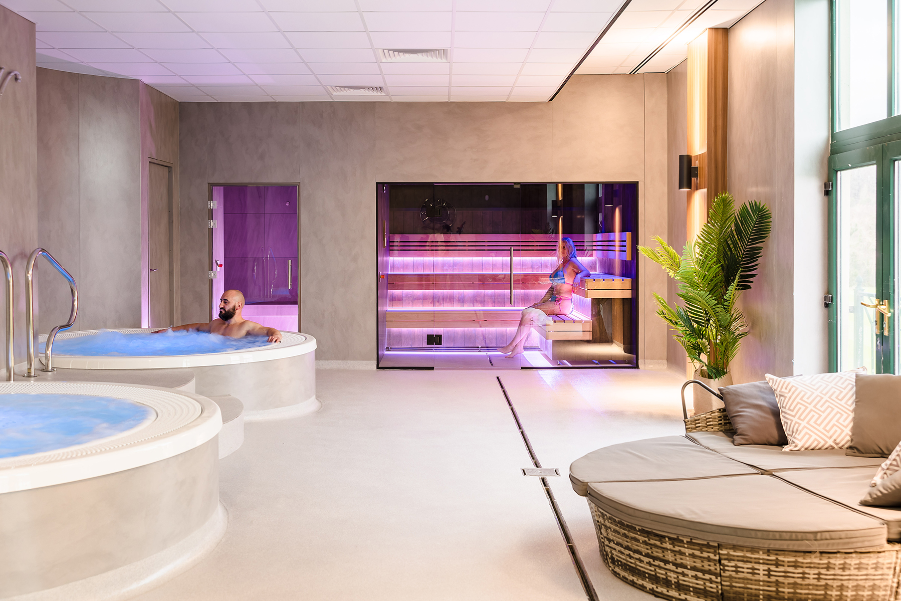Spa room with a man in a hot tub and a woman sitting in an infrared sauna. There's a lounge chair and a potted plant nearby.