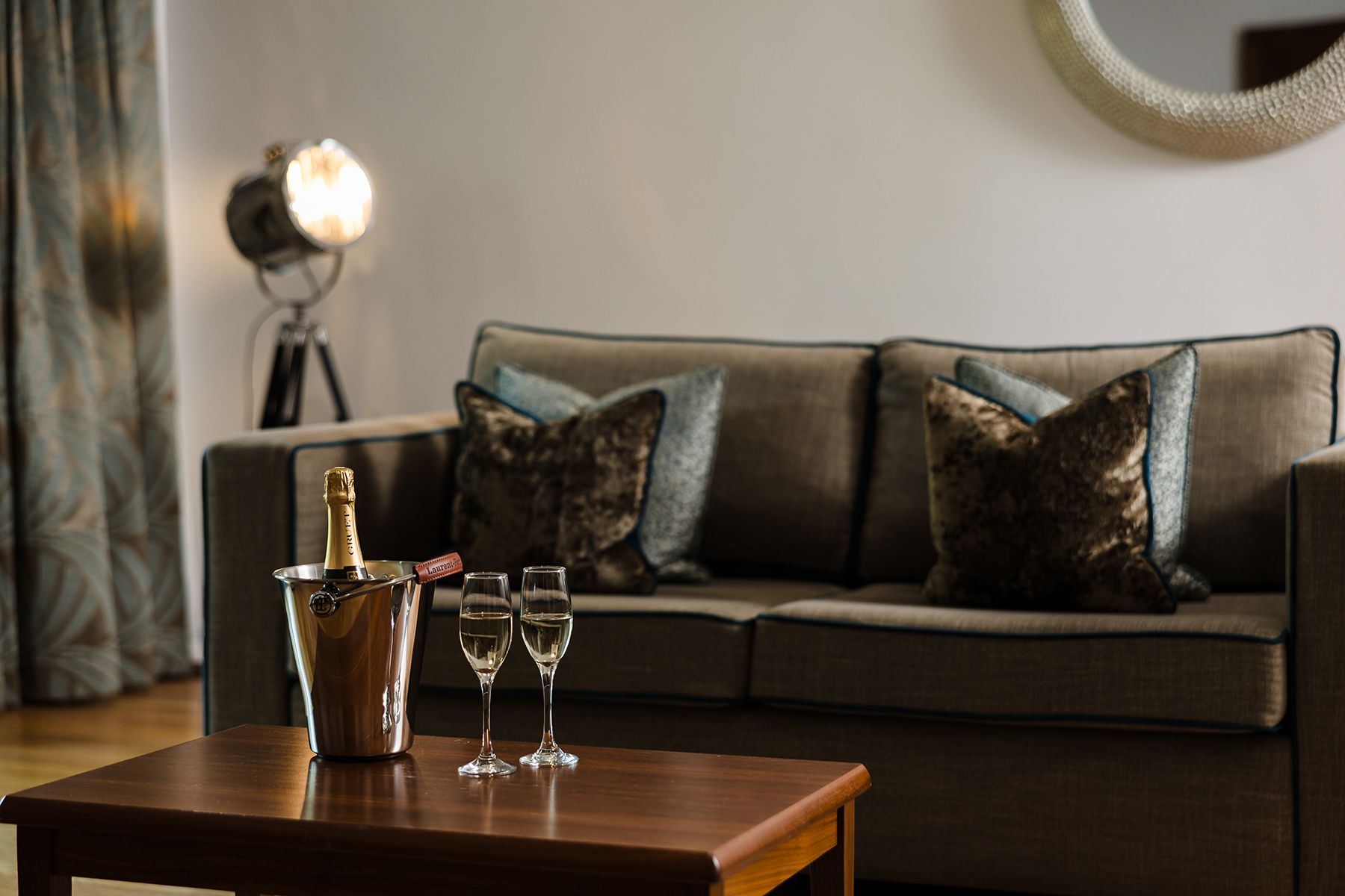 Two glasses of champagne and a bottle in an ice bucket on a wooden table in front of a gray sofa with cushions, next to a spotlight lamp and a circular mirror.