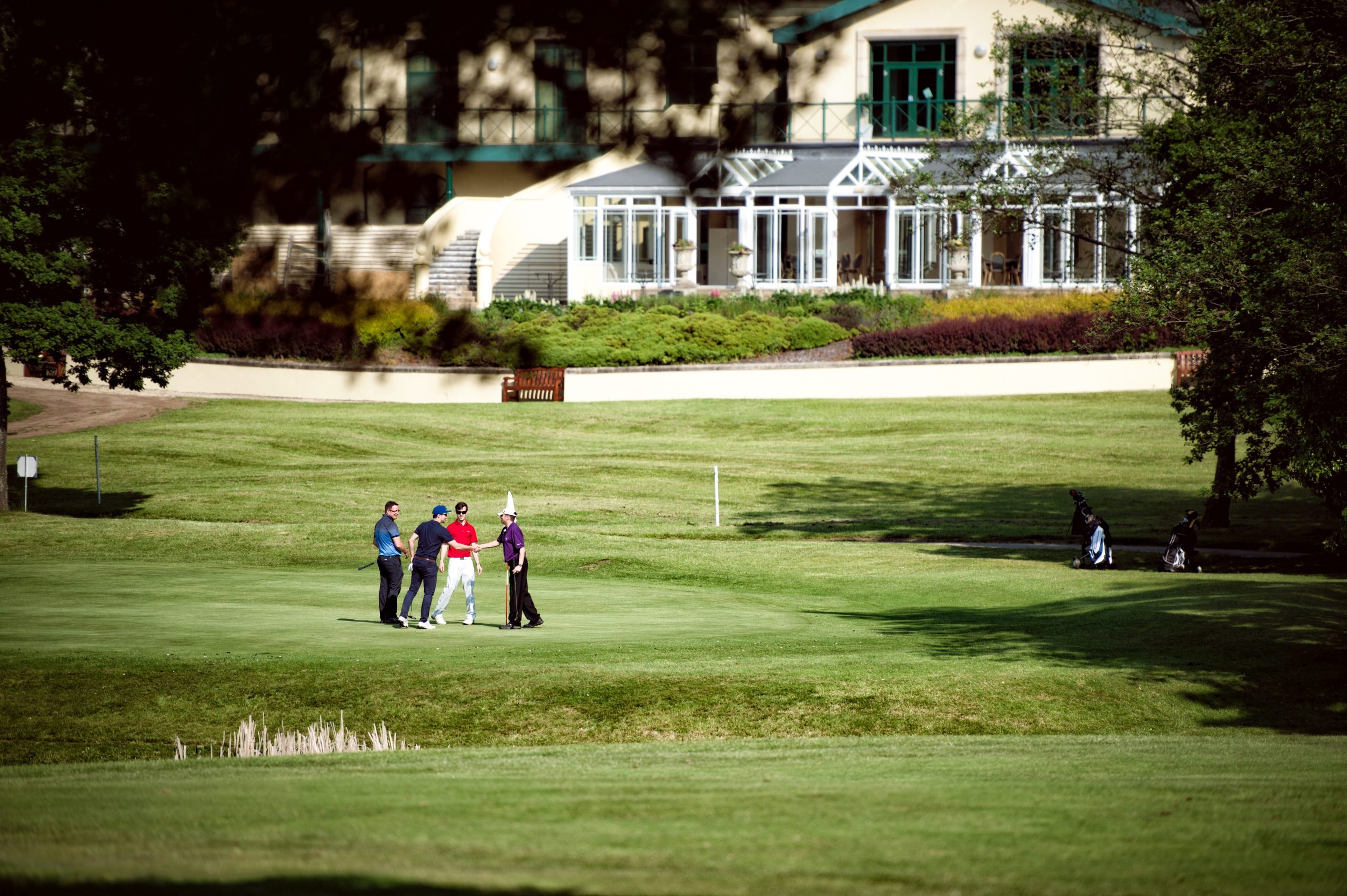 Four people stand on a golf course, shaking hands near a clubhouse in the background. Golf bags are visible on the grass.