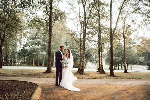 A bride and groom stand together on a forest path, surrounded by tall trees, in a serene outdoor setting.