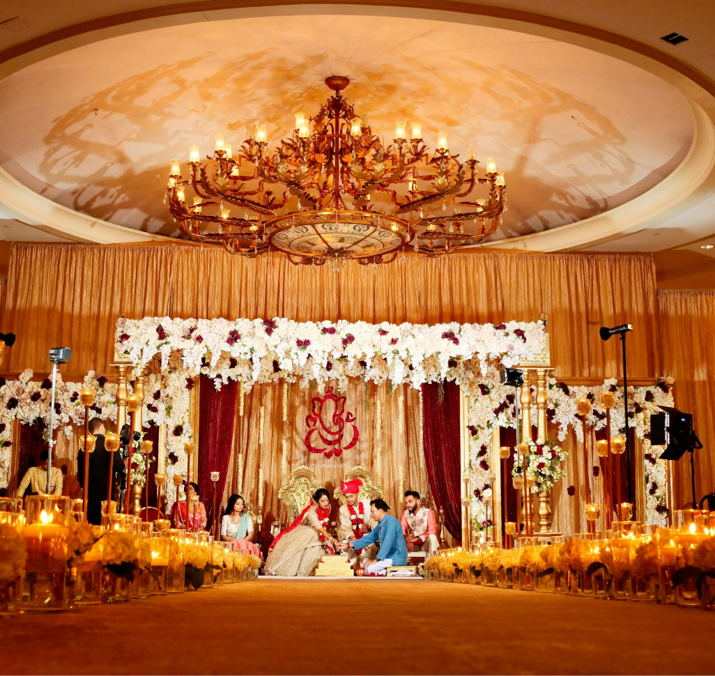 A couple sits under a large chandelier in a lavishly decorated venue with gold drapes and floral arrangements.