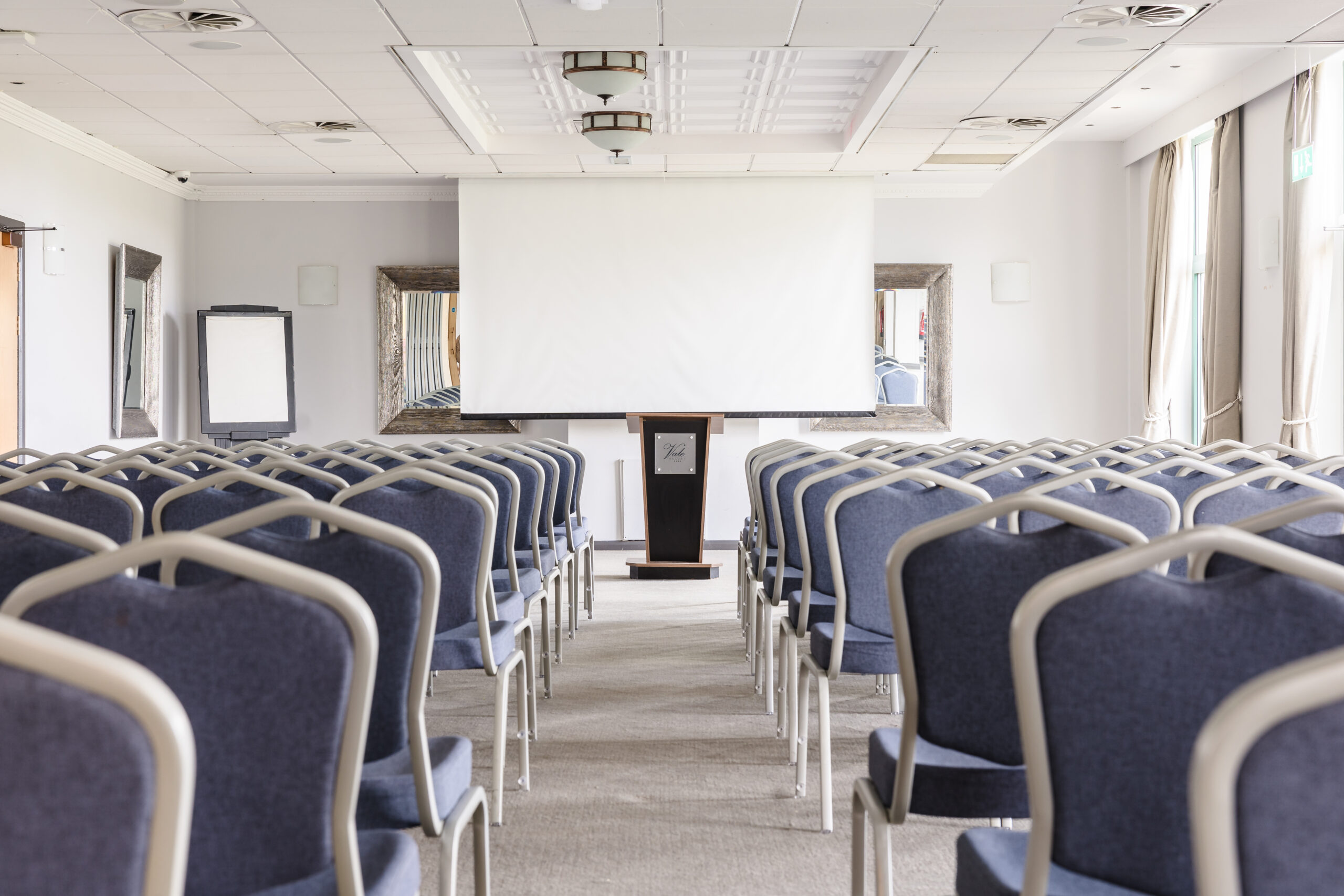 Empty conference room with rows of blue chairs facing a podium and projection screen.