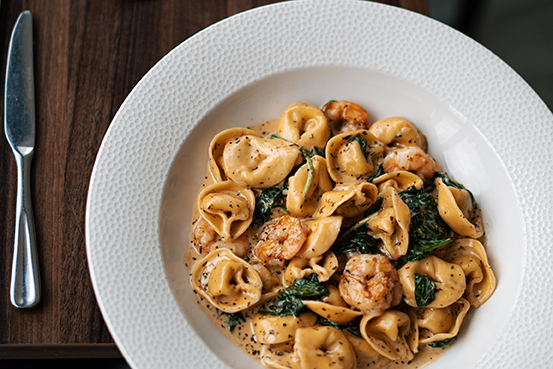 A bowl of tortellini with shrimp and greens in a creamy sauce on a wooden table, with a fork beside the plate.