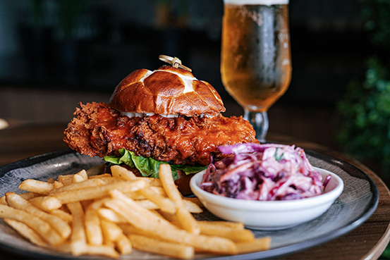 A plate with a fried chicken sandwich on a pretzel bun, served with fries and a side of coleslaw. A glass of beer is in the background.