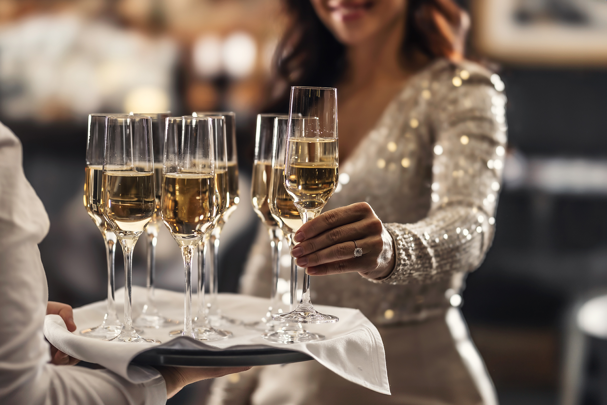 A person in a sequin dress takes a glass of champagne from a tray holding several other filled champagne glasses.