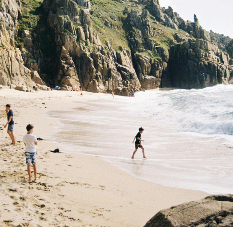 People walking and playing near the water on a sandy beach with rocky cliffs in the background.