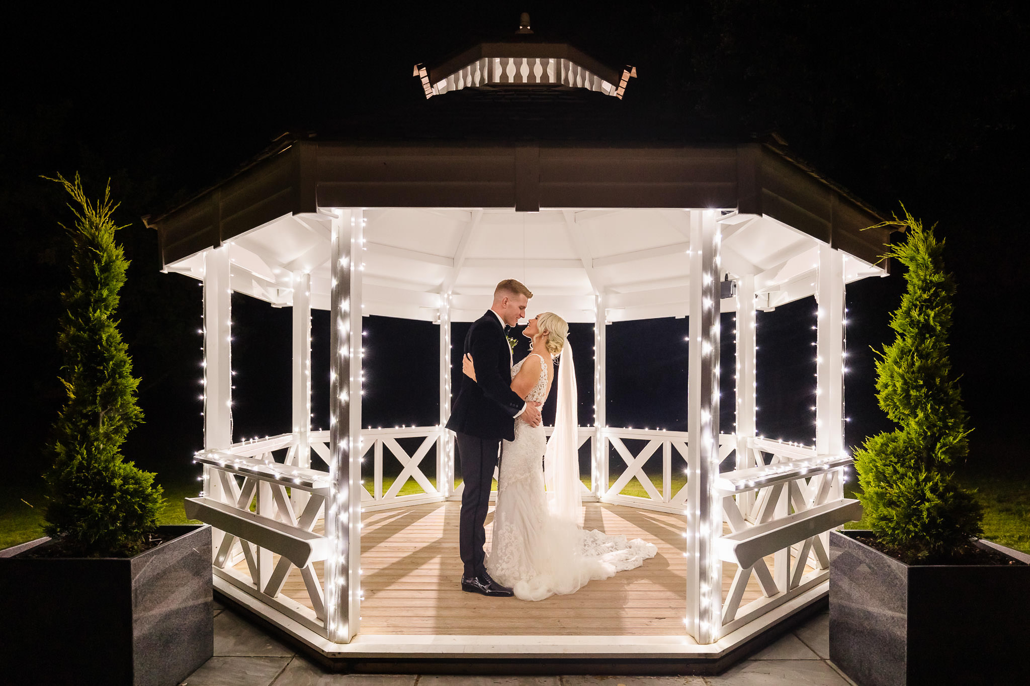 A couple stands in a lit gazebo at night, dressed in formal attire, surrounded by string lights.