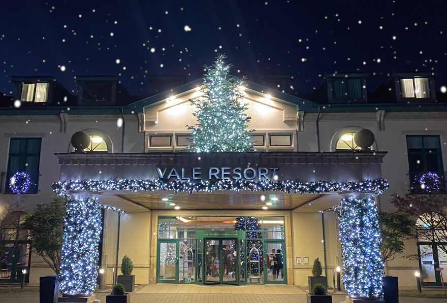 Entrance of Vale Resort decorated with festive lights and a tree. Snowflakes are falling in the evening sky.