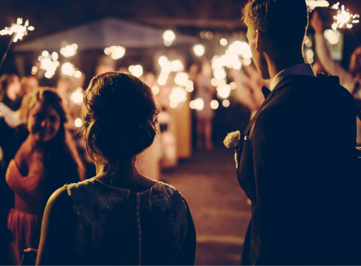 A couple stands facing a crowd of people holding sparklers at an evening event.