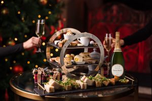 A festive table with afternoon tea snacks, a bottle of champagne, and two people toasting near a decorated Christmas tree.