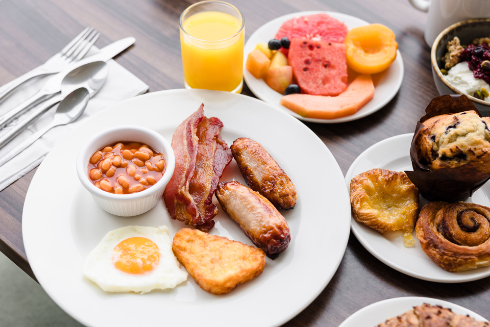 A breakfast plate with baked beans, bacon, sausages, hash brown, and a fried egg, with juice, fruit, and pastries nearby.