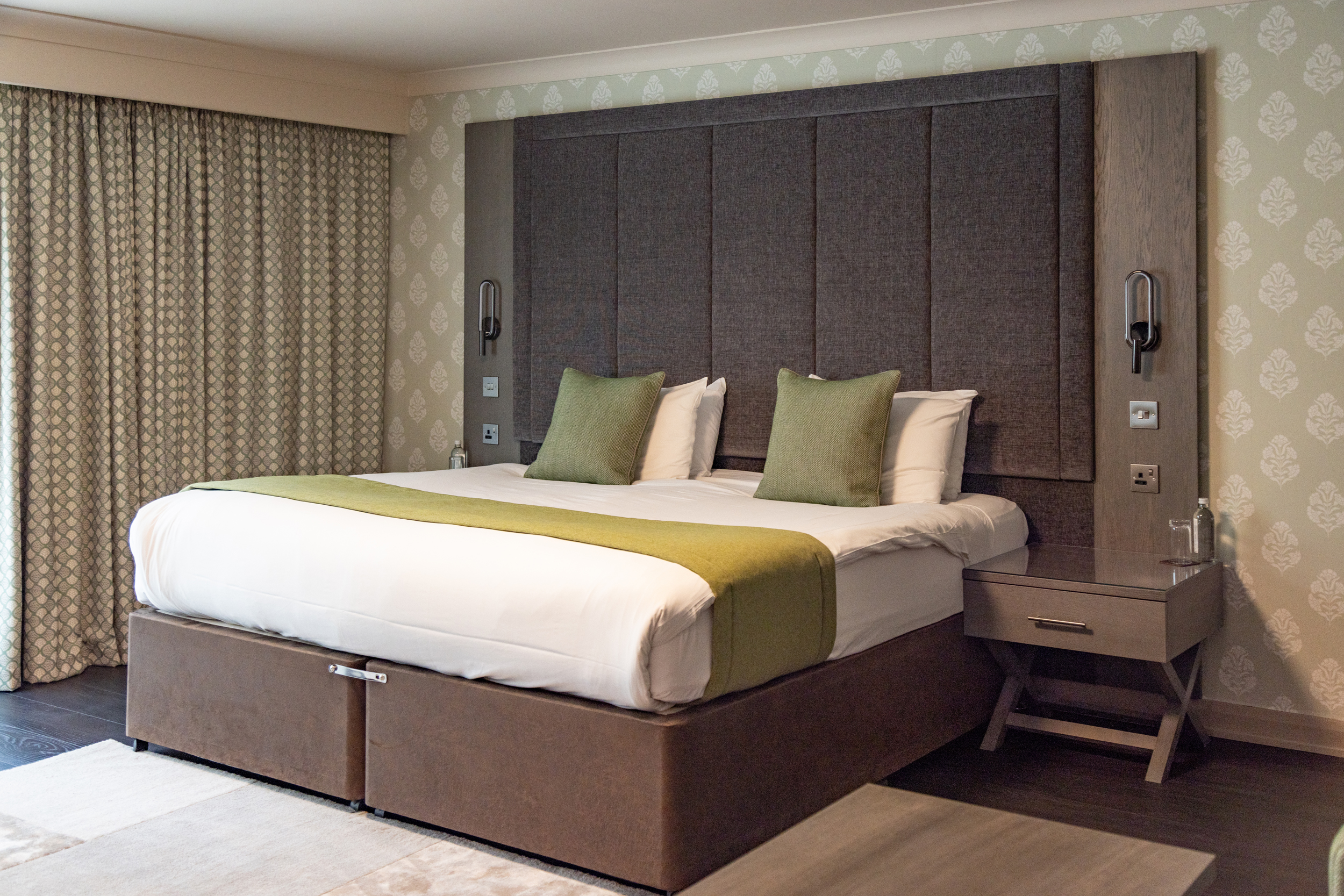 Modern hotel room with a king-sized bed, green accents, two pillows, and a bedside table with a lamp.