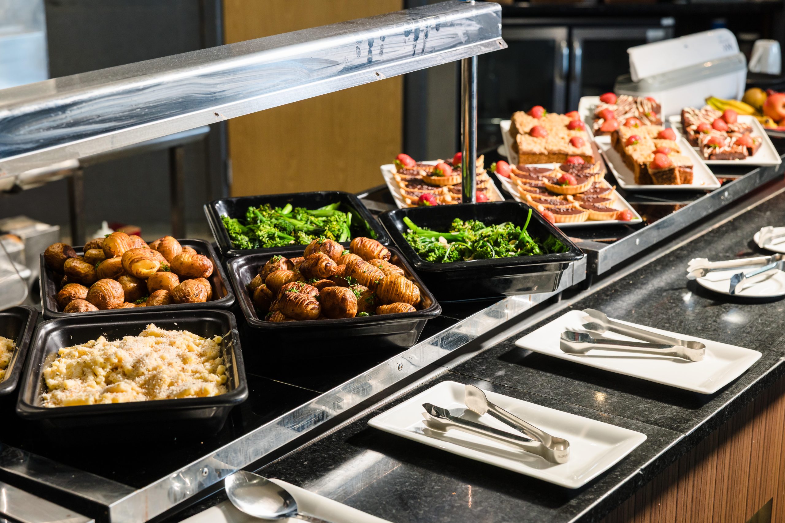 Buffet with trays of roasted potatoes, greens, rice, and various appetizers beside a stack of plates and cutlery.