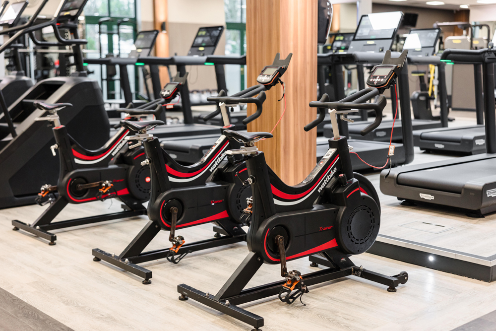 Three stationary exercise bikes are lined up in a gym, with treadmills visible in the background.