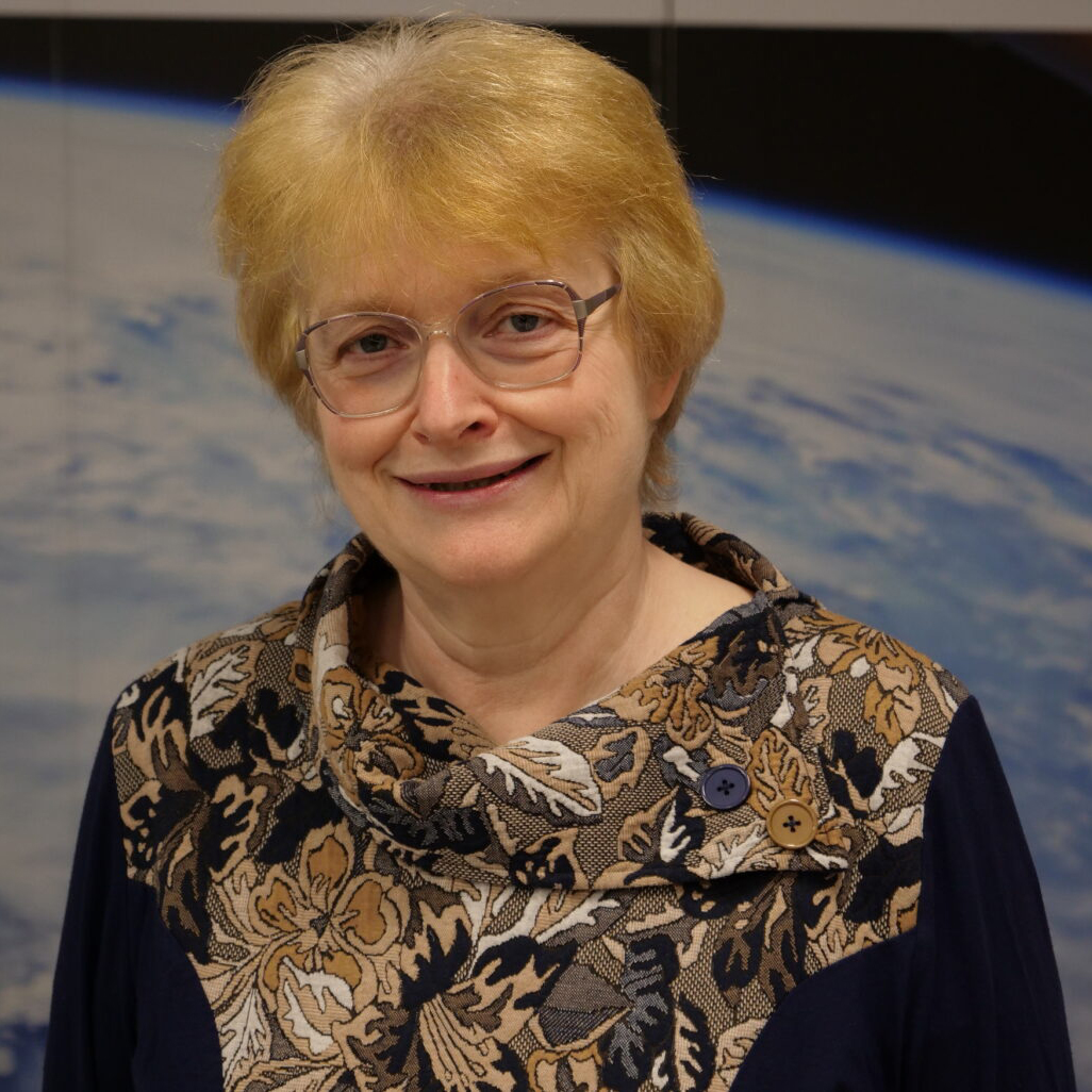 A woman with short blonde hair, glasses, and a patterned top stands in front of an image of Earth from space.