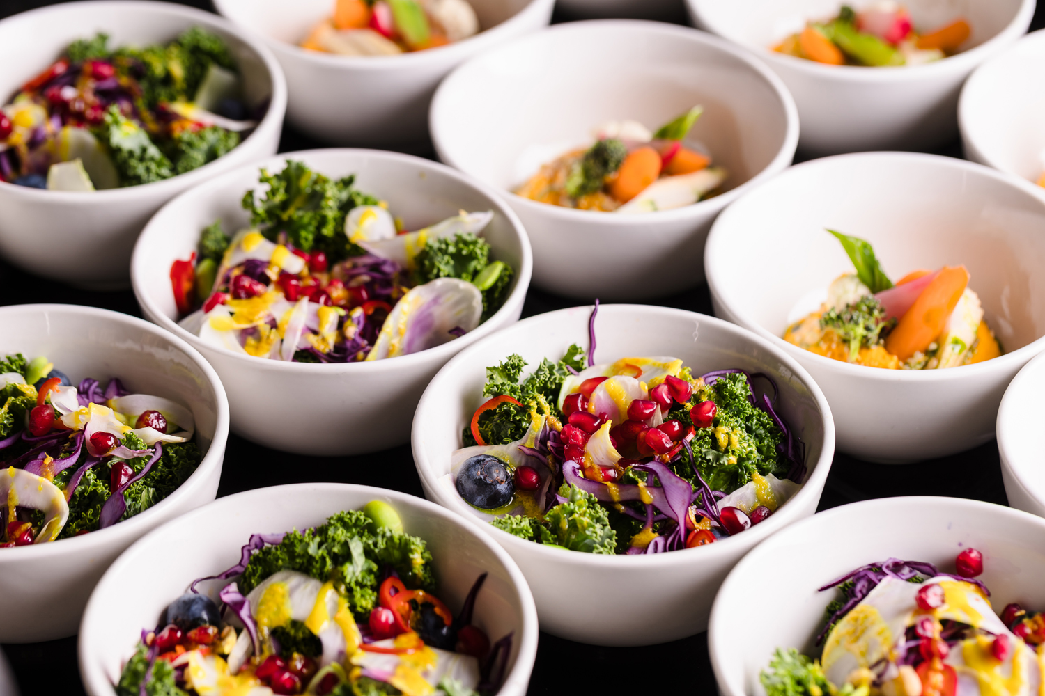 Bowls of colorful salads with kale, pomegranate seeds, and various vegetables, arranged in rows on a black surface.