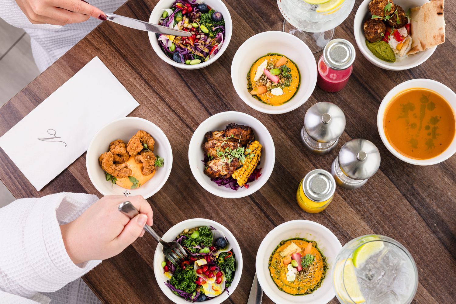 A wooden table set with various small bowls of colorful dishes, drinks, and condiments. Hands using cutlery visible.