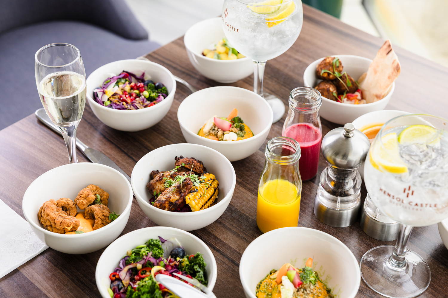 Assorted dishes in white bowls on a wooden table with glasses of drinks, including wine and water with lemon slices.