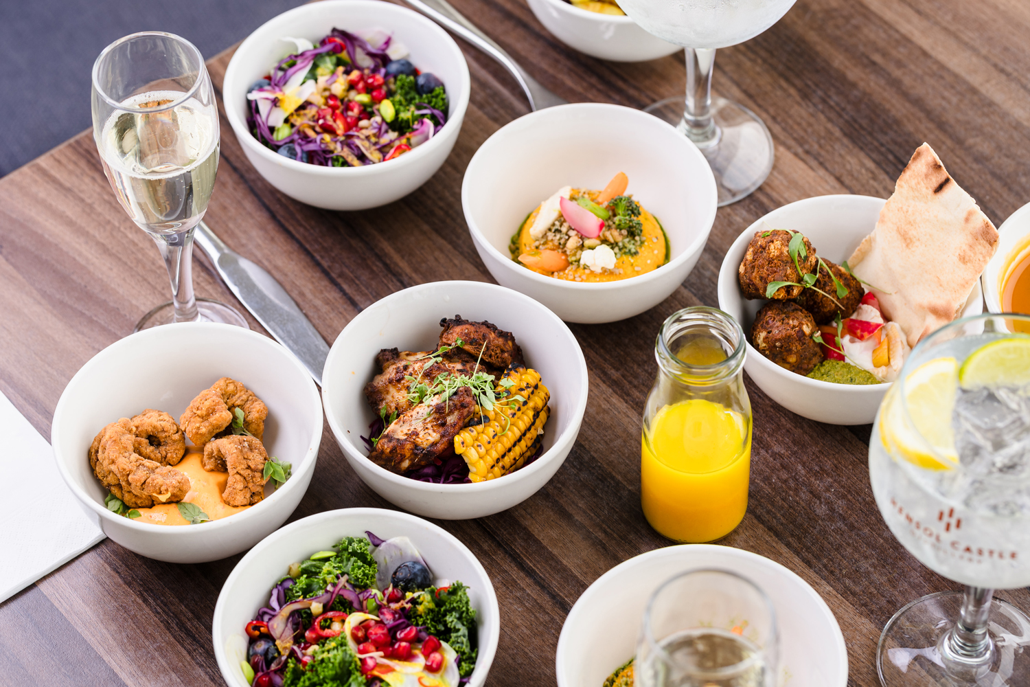 A variety of dishes in white bowls on a wooden table, accompanied by glasses of drinks, including juice and sparkling wine.