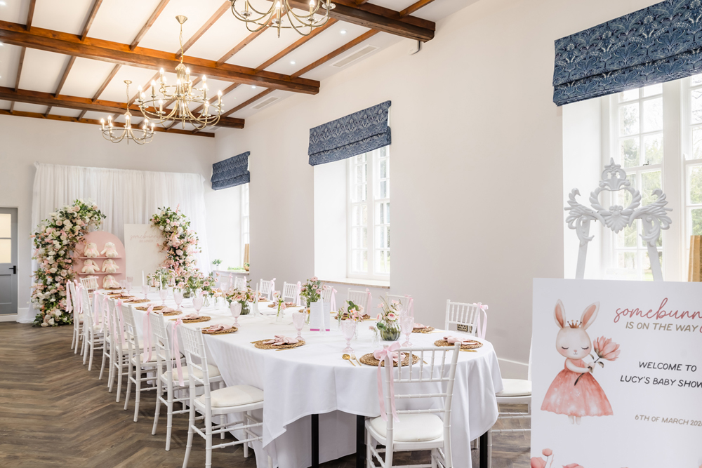 Elegant baby shower setup with a large table, pink floral decorations, and a sign with a bunny illustration in a bright room.