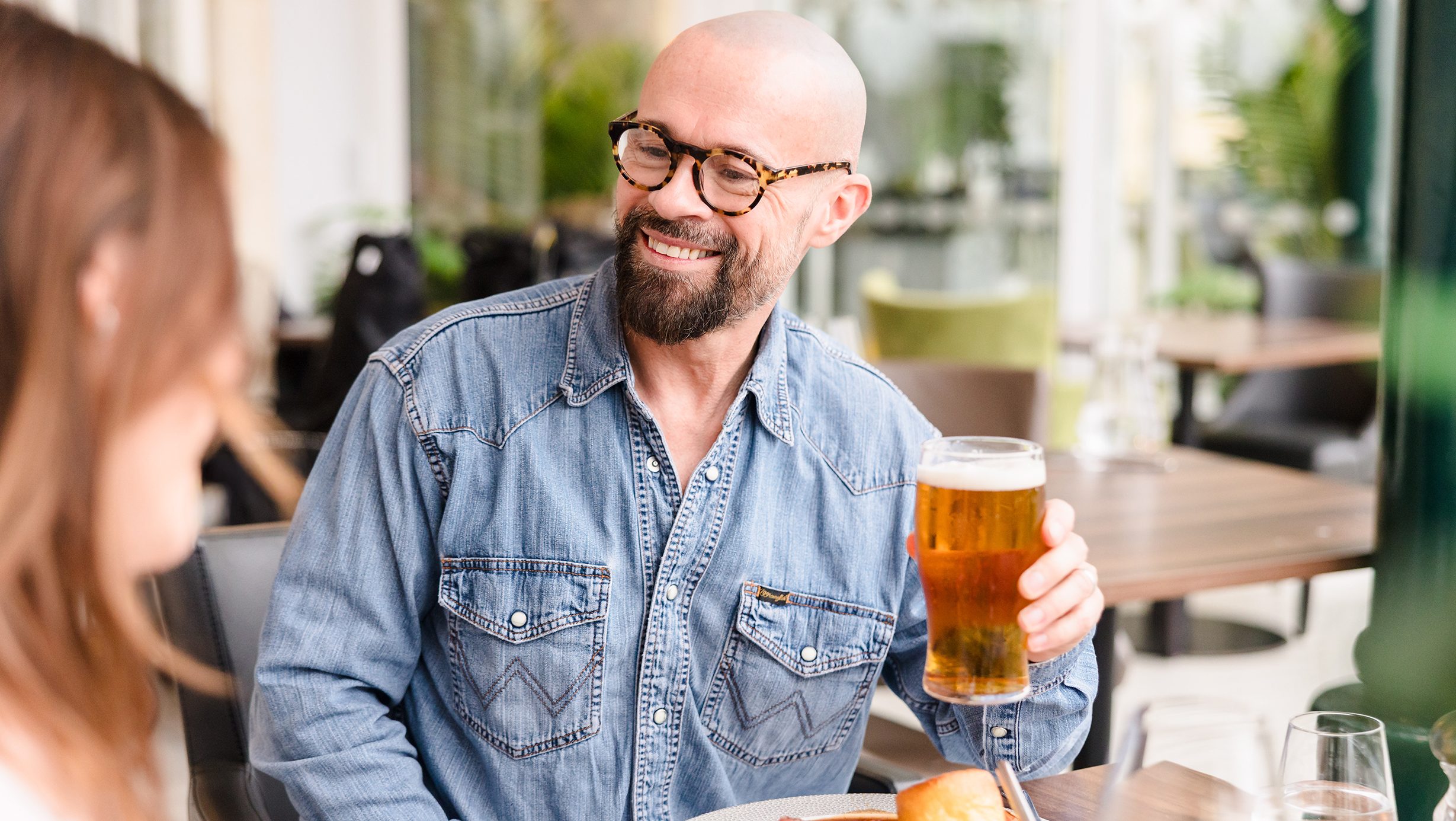 A man in glasses and a denim shirt holds a pint of beer while sitting at a restaurant table with food.