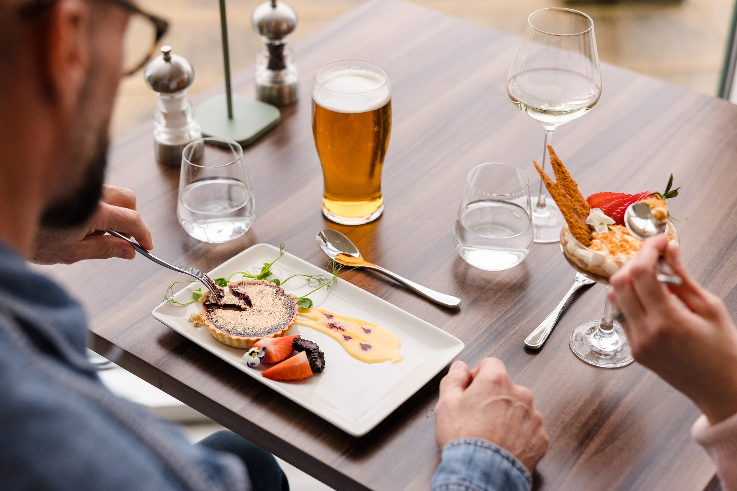 Two people dine at a table with plated desserts, a glass of beer, wine, and water glasses on a wooden surface.