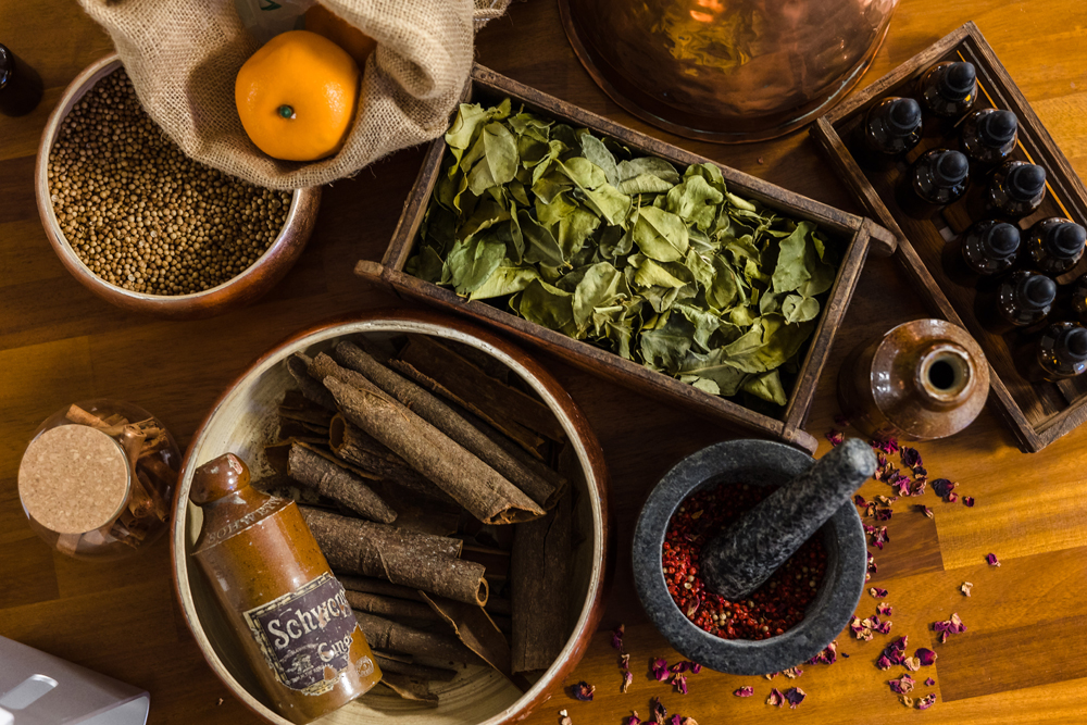 Variety of herbs and spices on a wooden table, including cinnamon sticks, dried leaves, a mortar with pestle, and small bottles.