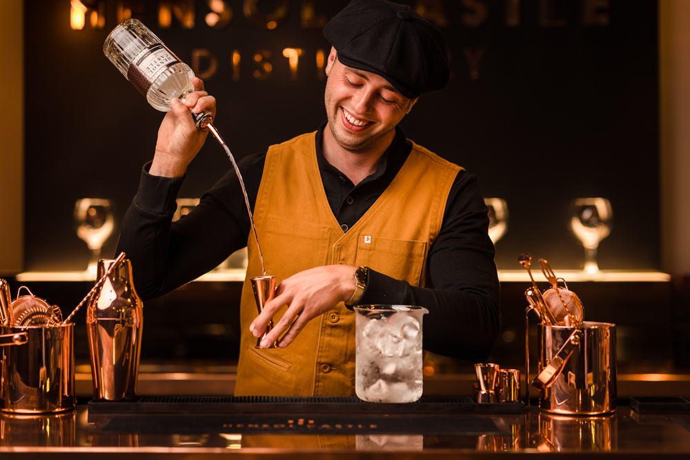 Bartender in a vest pours alcohol into a jigger over a glass of ice at a bar with copper equipment.