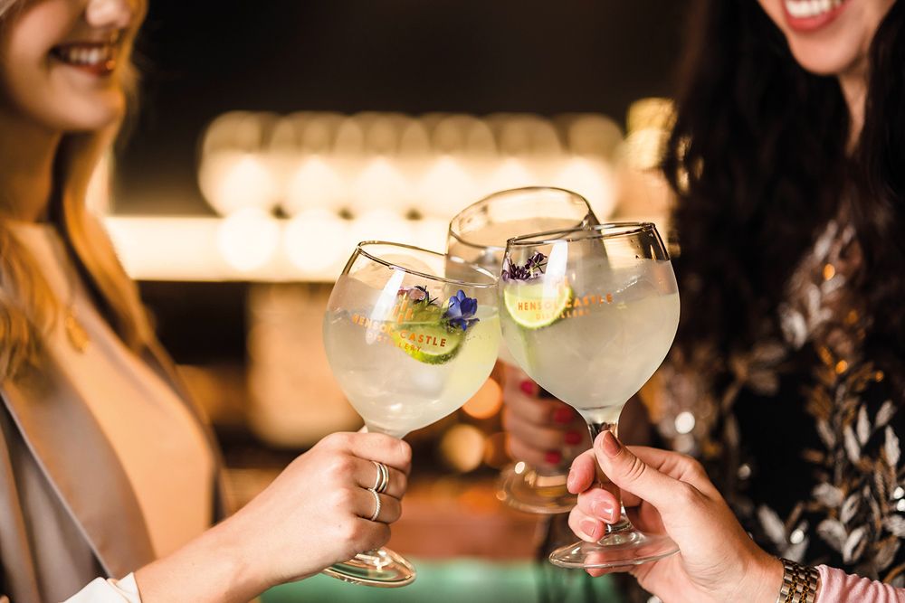 Three people clinking large cocktail glasses with lime and flowers, celebrating in a dimly lit setting.