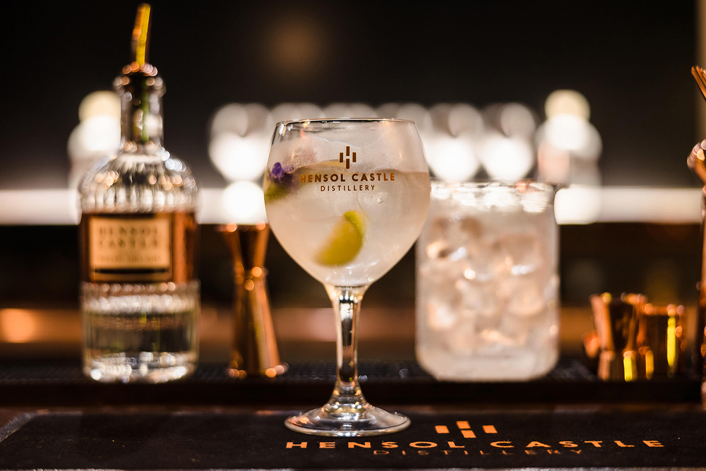 A glass with Hensol Castle logo contains a drink and a lemon slice; bottle and ice-filled glass blurred in the background.