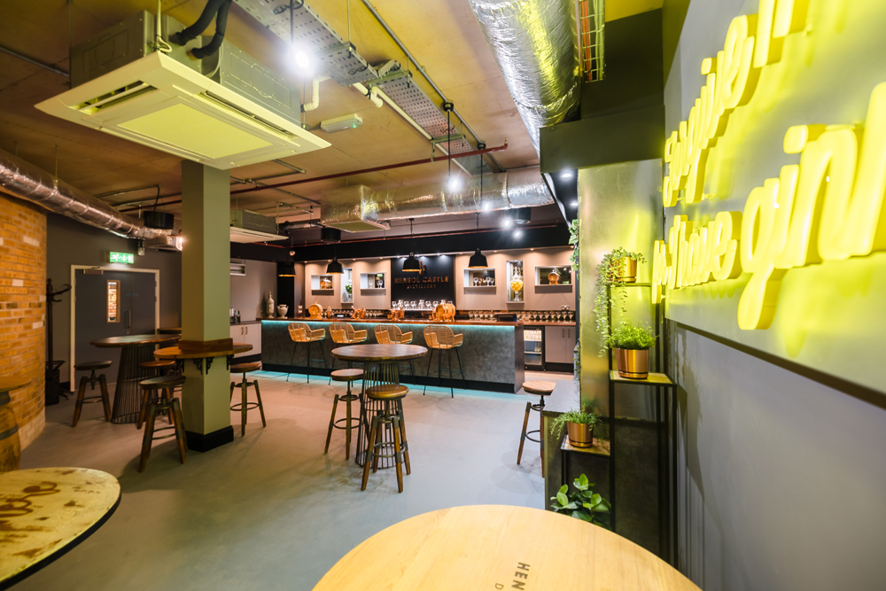 Modern bar with high stools, round tables, exposed ductwork, and neon sign on the wall. Ceiling lights illuminate the space.