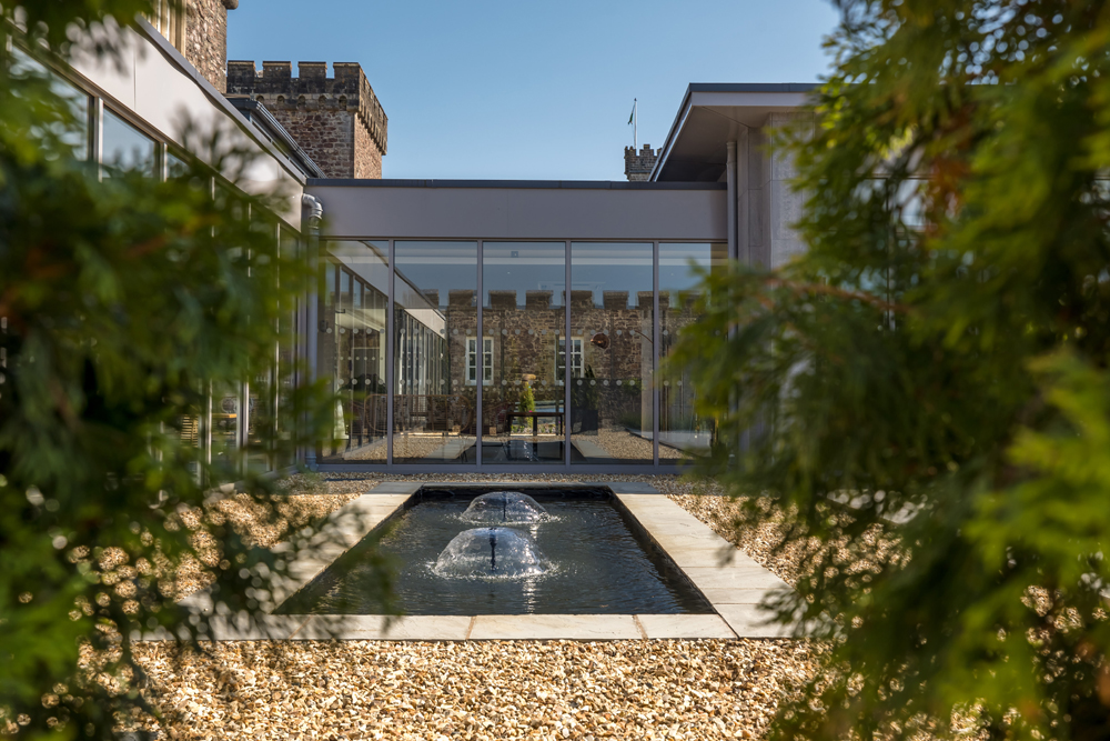 View of a modern building with glass walls, a stone courtyard, and a central water fountain, framed by greenery.