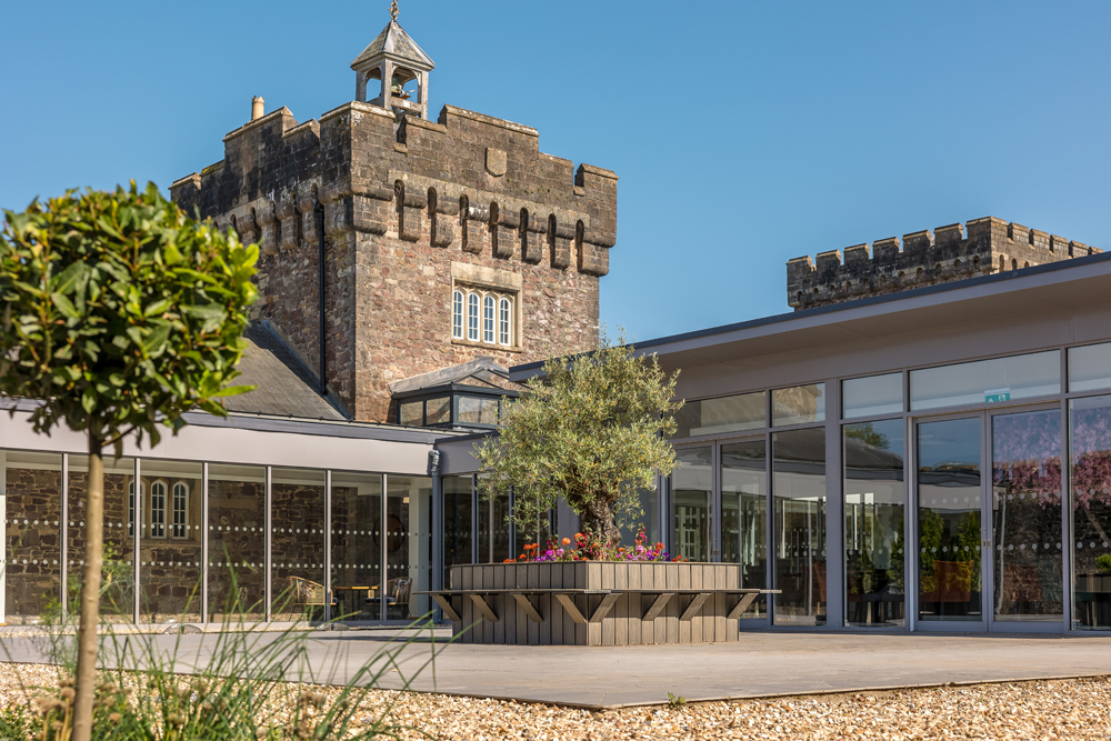 Modern glass extension with planter against a historic stone castle backdrop under a clear blue sky.