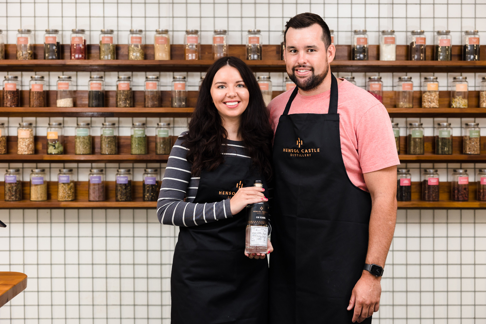 Two people in aprons stand together in front of shelves with jars, holding a bottle.