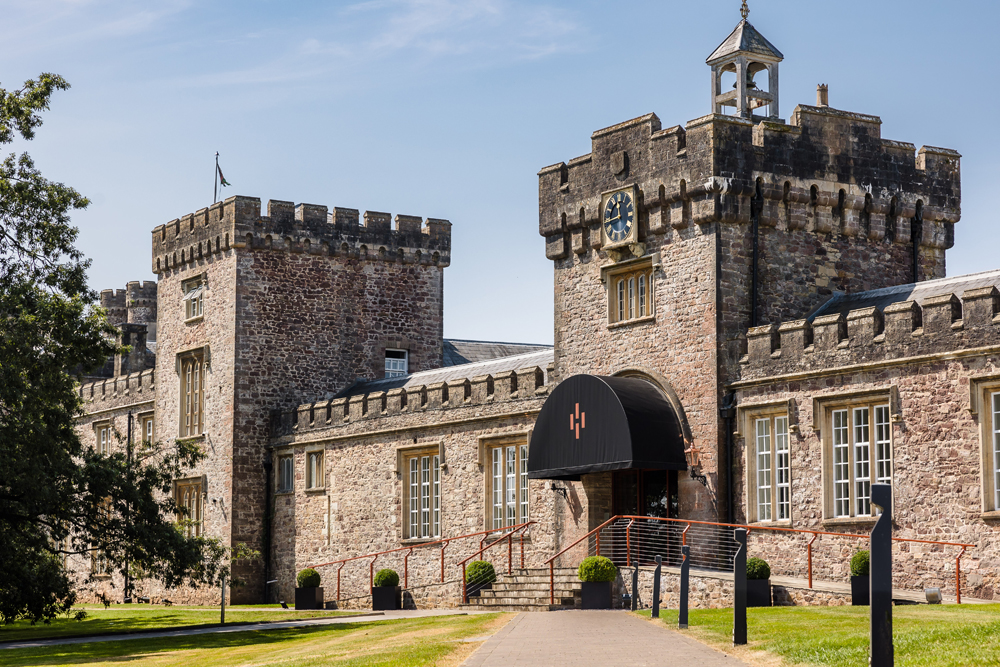 Stone castle with crenellated towers, arched entrance, and a clock, set in a grassy area with a clear blue sky.