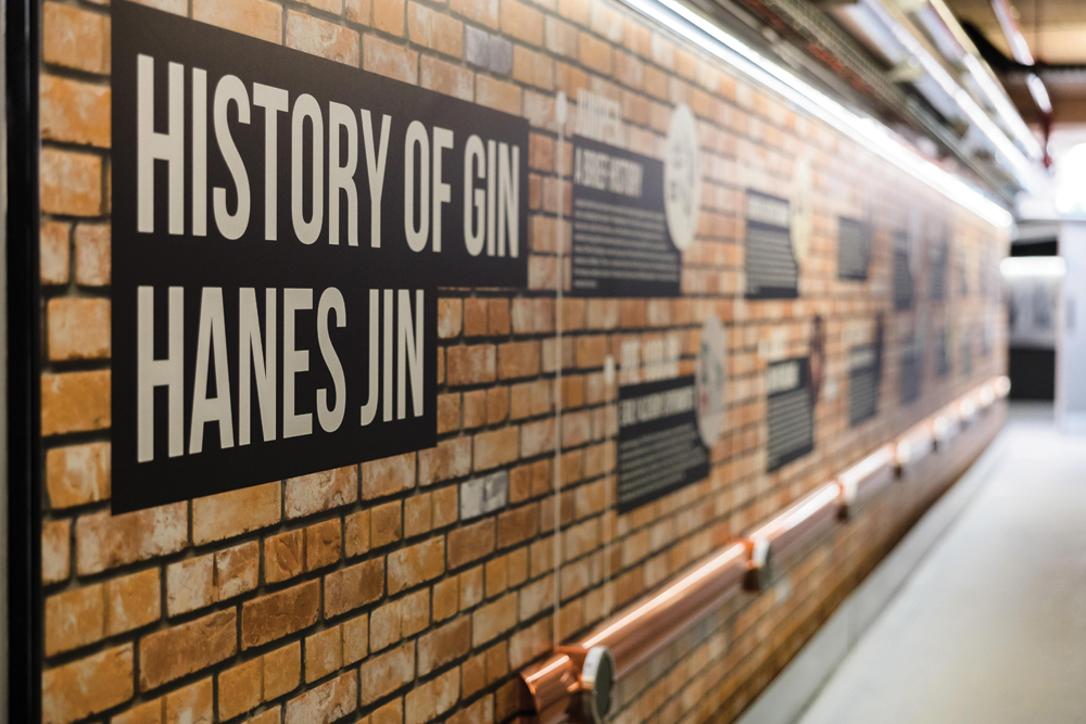 Brick wall display with text panels about the history of gin, labeled "Hanes Jin," in a museum or exhibition setting.