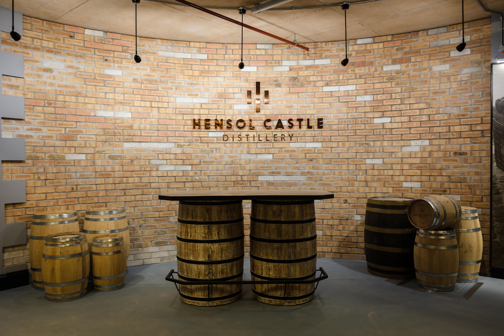 Room with a brick wall, a table made of barrels, and additional barrels beside it at Hensol Castle Distillery.